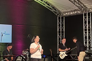 Eine Person steht auf einer Bühne und spricht in ein Mikrofon. Im Hintergrund sind drei Musiker mit Instrumenten: ein Schlagzeuger, ein Gitarrist und ein Bassist. Die Bühne ist mit bunten Lichtern beleuchtet.