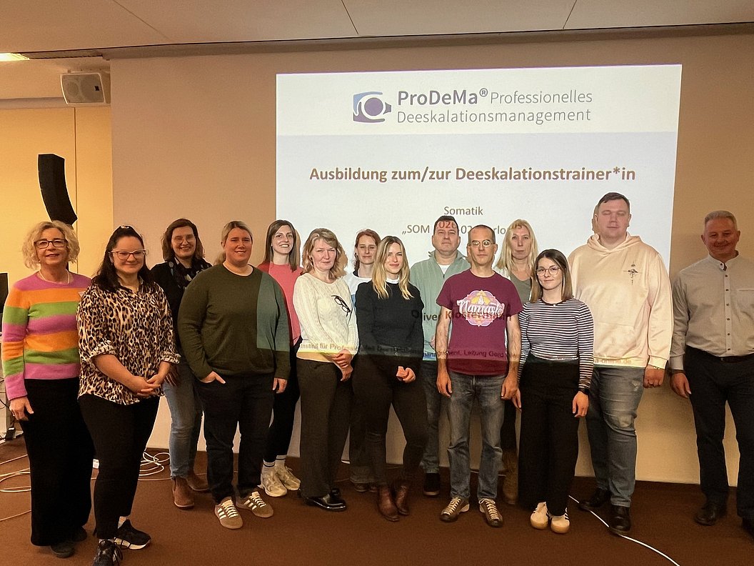Gruppe von Menschen steht vor einer Projektionswand mit dem Text 'ProDeMa Professionelles Deeskalationsmanagement Ausbildung zum/zur Deeskalationstrainer*in'