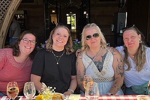 Vier Frauen sitzen an einem rot-weiß karierten Tisch mit Gläsern und Blumen in einem Restaurant.