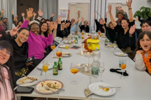 Gruppe von Menschen sitzt an einem langen Tisch mit Tellern und Getränken, viele heben die Hände und zeigen Friedenszeichen.