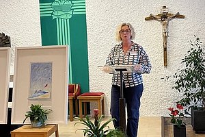Frau steht an einem Rednerpult in einer Kirche mit Kreuz an der Wand und mehreren Pflanzen im Vordergrund