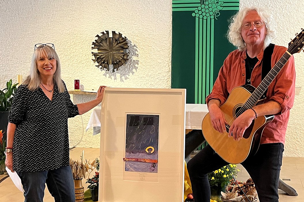 Zwei Personen in einem Raum, eine hält einen gerahmten Kunstdruck, die andere sitzt mit einer Gitarre auf einem Stuhl, im Hintergrund ein grünes Banner und eine Wanddekoration.