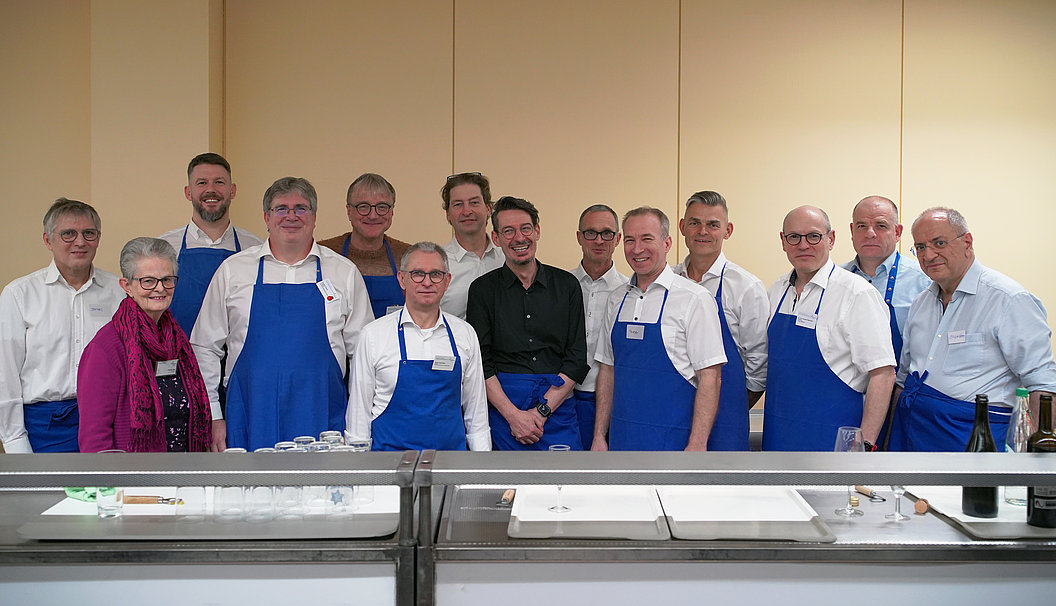 Gruppe von Menschen in weißem Hemd und blauer Schürze steht hinter einem langen Tisch mit Tabletts und Besteck in einem Raum mit beiger Wand