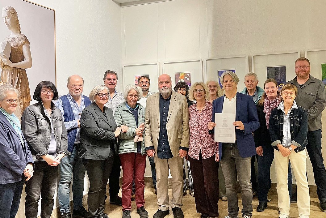 Gruppe von Menschen in einem Ausstellungsraum mit gerahmten Bildern an der Wand, eine Person hält ein Dokument in der Hand.