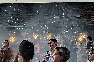 Menschen sitzen an Tischen mit karierten Tischdecken im Freien vor einem Gebäude mit Dachfenstern bei Sonnenuntergang