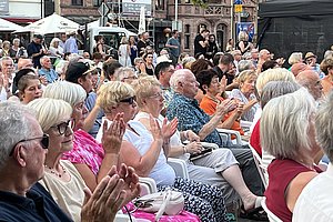 Eine Menschenmenge sitzt auf weißen Stühlen und applaudiert. Die Zuschauer sind überwiegend älter, tragen verschiedene Kleidung und sitzen in einem Stadtbereich mit historischen Gebäuden im Hintergrund.
