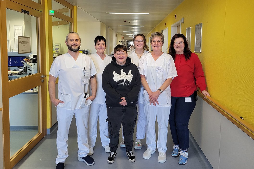 Fünf Personen stehen in einem Krankenhausflur, vier tragen weiße medizinische Kleidung, eine Person trägt eine rote Jacke und blaue Schuhe.