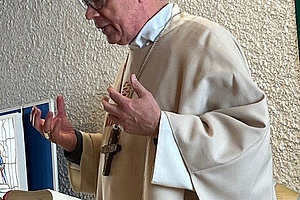 Bischof Ackermann mit rosa Birett und cremefarbener liturgischer Kleidung steht an einem Lesepult mit aufgeschlagenem Buch und hebt die Hände.