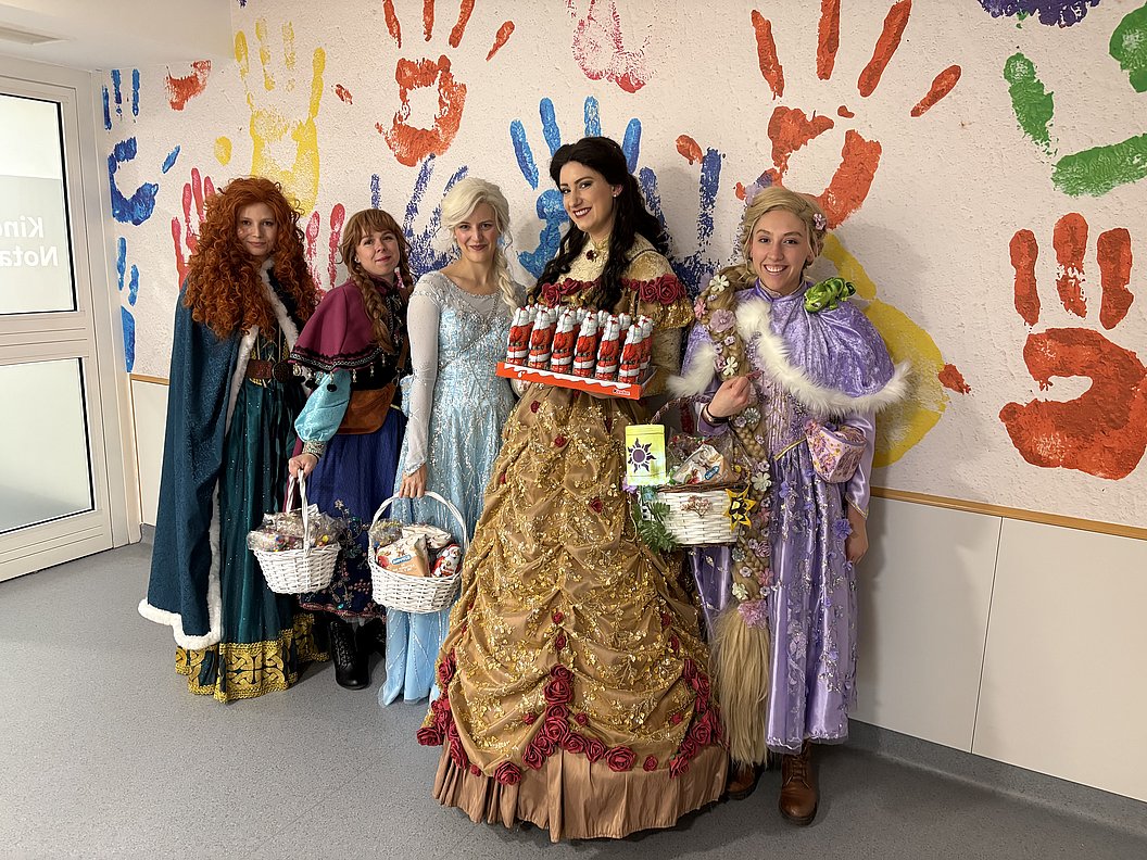 Fünf Prinzessinnen in Disney-Kostümen mit Körben stehen vor einer Wand mit bunten Handabdrücken.