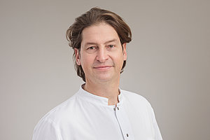 Chefarzt Dr. Joachim Wagner trägt einen weißen Arztkittel und steht vor einem neutralen Hintergrund
