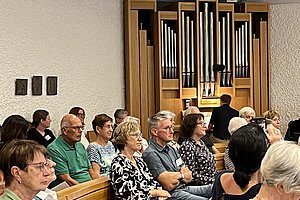 Eine Gruppe von Personen sitzt in einer Kirche oder einem Veranstaltungsraum. Im Hintergrund steht eine große Orgel.