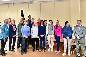 Gruppe von Menschen unterschiedlichen Alters und Geschlechts, stehend in einem Raum mit hellen Vorhängen und Stühlen im Hintergrund.