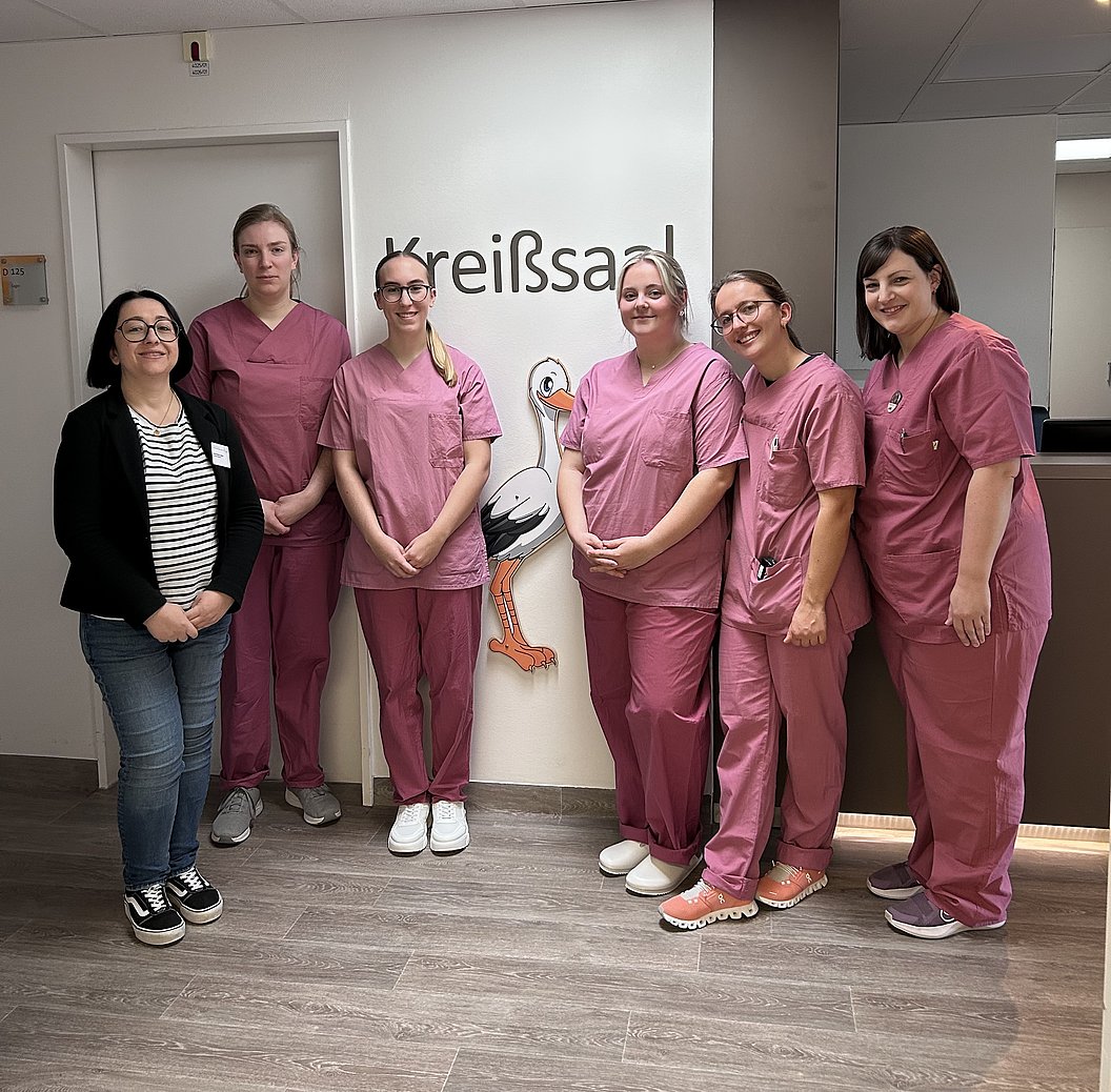 Sechs Personen, fünf in rosa medizinischer Kleidung, eine in Freizeitkleidung, stehen vor einer Tür mit der Aufschrift 'Kreißsaal' und einer Storchillustration.