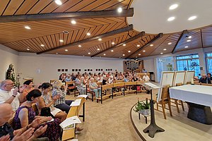 Menschen in einer Kapelle mit Holzdecke, Orgel und Altar, Teilnehmer sitzen auf Bänken und applaudieren