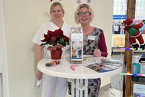 Zwei Frauen mit weihnachtlichen Haarreifen stehen an einem Stehtisch mit Weihnachtsstern, Flyern und einer Spendenbox in einem hellen Raum