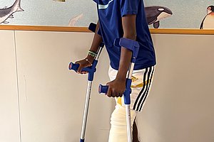 Ein Junge mit einem Gipsverband am linken Bein steht auf Krücken und hält einen Fußball. Er trägt ein blaues T-Shirt und eine Sporthose mit Streifen.