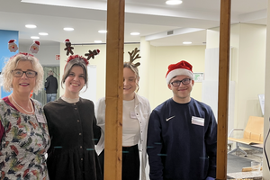 Vier Personen stehen in einem Raum, zwei tragen Weihnachtskopfbedeckungen, vor ihnen steht ein Tisch mit Tellern und Servietten