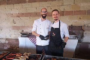 Zwei Köche in weißer Kleidung und schwarzer Schürze stehen an einem Grill mit verschiedenen Würstchen und Burgern vor einer Backsteinwand.