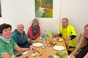 Fünf Personen sitzen um einen rechteckigen Holztisch mit Wasserflaschen, Obst, Salat und Brot in einem Raum mit weißer Wand und Landschaftsgemälde.