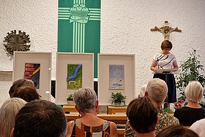 Frau steht am Rednerpult in einer Kirche mit Kreuz und grünem Banner, Publikum sitzt auf Holzbänken