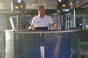 Person steht hinter einem DJ-Pult mit LED-Beleuchtung unter einem Zelt mit Lautsprechern und Lichttechnik