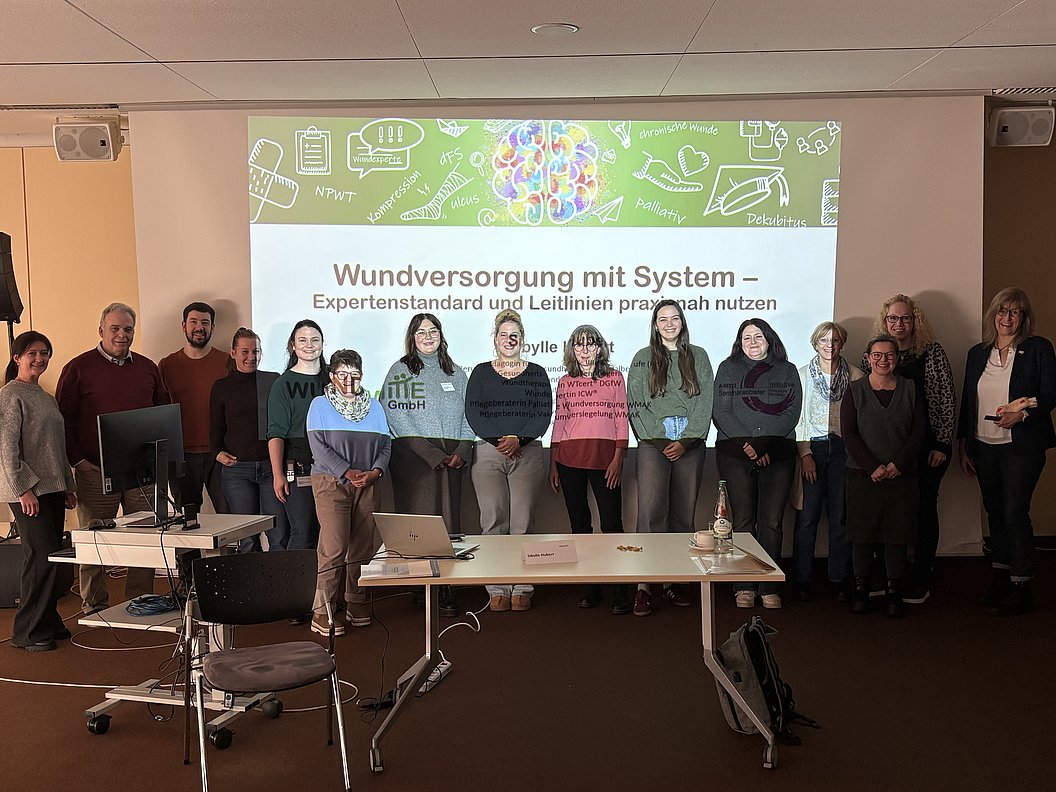 Gruppe von Menschen steht vor einer Projektionswand mit Text 'Wundversorgung mit System – Expertenstandard und Leitlinien praxisnah nutzen'.