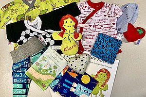 Mehrere bunte Kinderkleidungsstücke und Stoffbücher liegen auf weißem Untergrund verteilt.