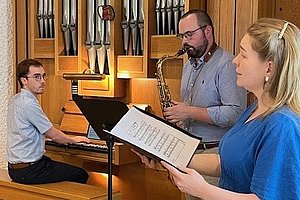 Drei Personen musizieren in einem Raum mit einer großen Orgel. Eine Person spielt Orgel, eine spielt Saxophon, eine singt mit Notenblatt in der Hand.