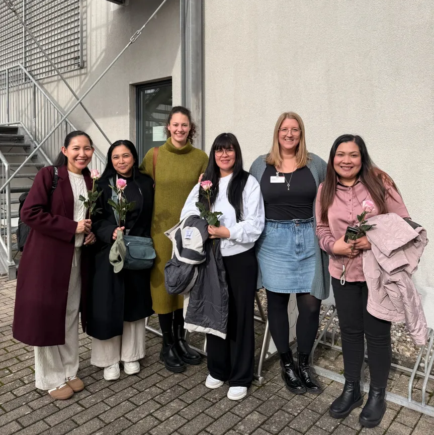 Sechs Personen stehen vor einer hellen Hauswand mit dem Logo 'MARIENHAUS'. Einige halten Rosen, eine trägt einen langen dunkelroten Mantel, eine andere einen Jeansrock.