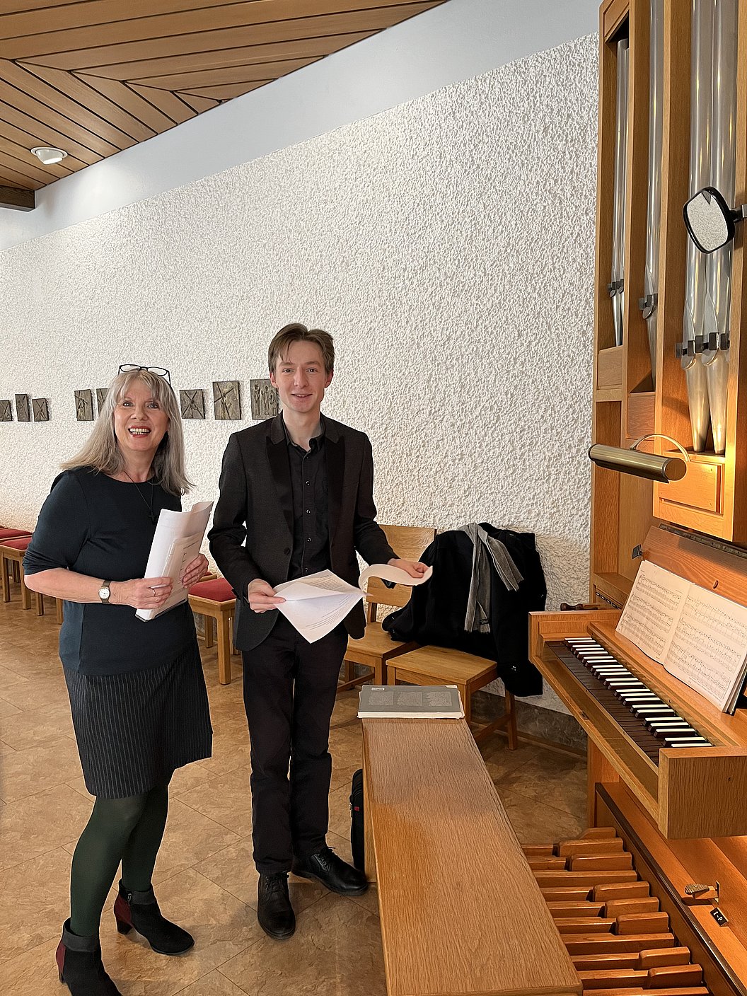 Zwei Personen stehen vor einer Orgel, beide halten Notenblätter in den Händen