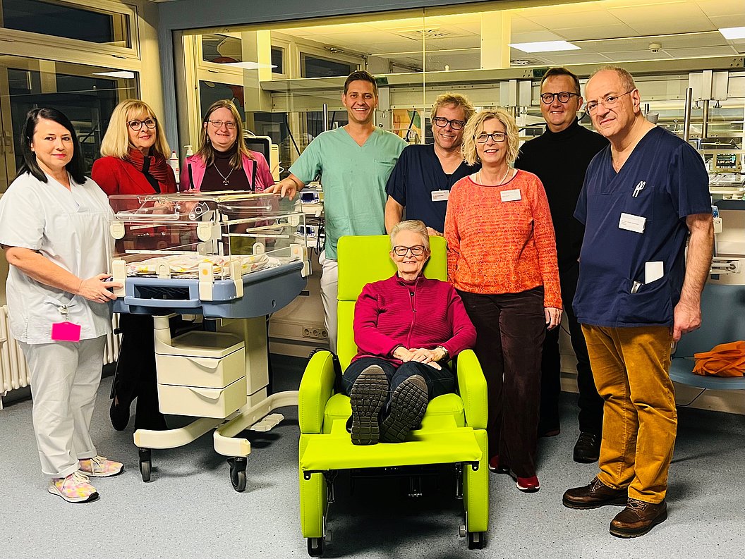 Gruppe von acht Personen in medizinischer Umgebung, eine Person sitzt in einem hellgrünen Stillsessel, daneben ein Inkubator für Neugeborene.