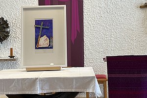 Altar mit weißem Tuch, darauf ein gerahmtes Bild mit Kreuz und Herz, im Hintergrund ein violettes Wandbanner mit stilisiertem Kreuz und Pfeilen