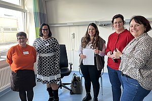 Fünf Frauen stehen in einem hellen Raum mit Stühlen und einem Fenster, einige halten Papiere in der Hand