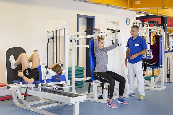Zwei Frauen trainieren an Fitnessgeräten, ein Mann in Sportkleidung steht daneben und eine weitere Frau beobachtet im Hintergrund