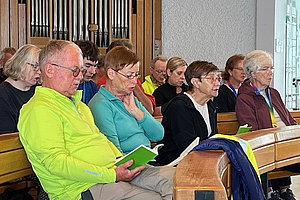 Mehrere Personen sitzen auf Holzbänken in einem Raum mit Orgelpfeifen im Hintergrund, einige halten grüne Hefte.