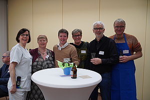 Sechs Personen stehen um einen hohen Stehtisch mit einer Bierflasche, einem Glas und einer blauen Vase mit grünen Servietten vor einer hellen Wand.