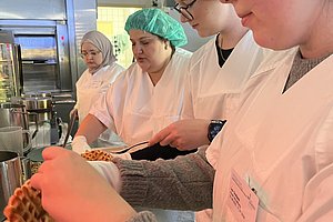 Mehrere Personen in Schutzkleidung und Haarnetzen bereiten in einer Küche Waffeln zu, eine Person hält eine Waffel über ein Waffeleisen