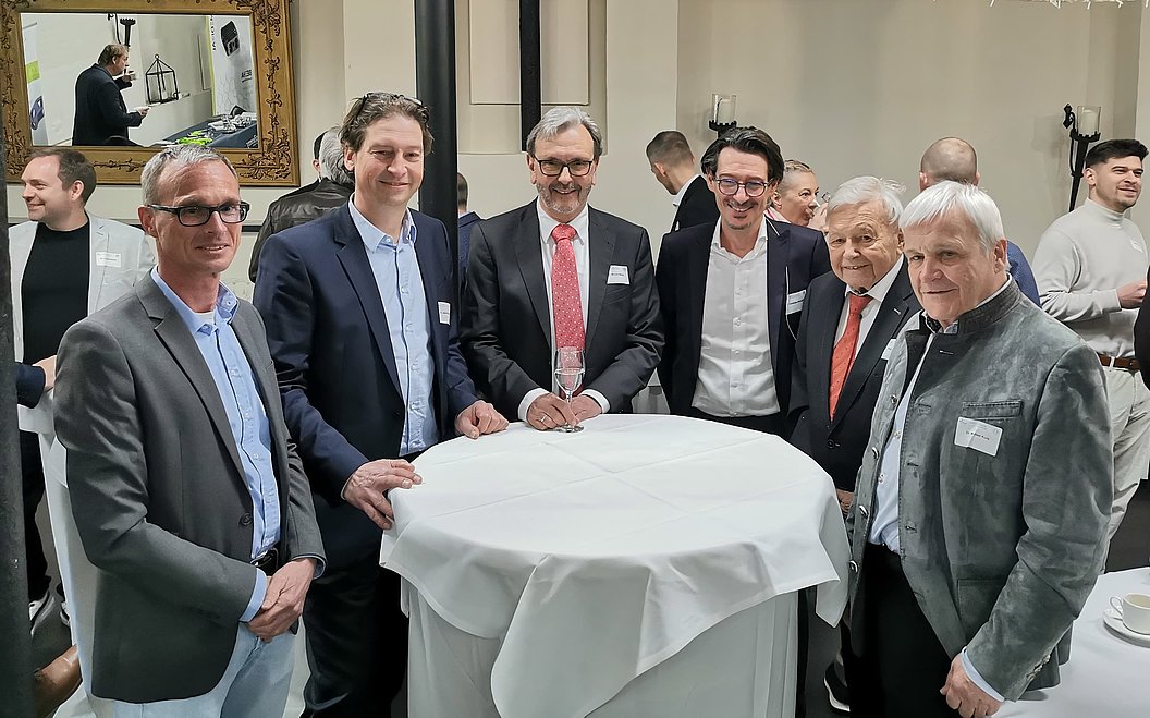 Sechs Männer in Anzügen stehen um einen mit weißem Tuch bedeckten Stehtisch in einem Konferenzraum