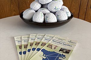 Schwarze Schale mit weißen Stressbällen, darauf gestapelt, und davor fünf Flyer mit Karte und Text 'NeuroNet SaarPlus' auf einem weißen Tisch vor Holzvertäfelung