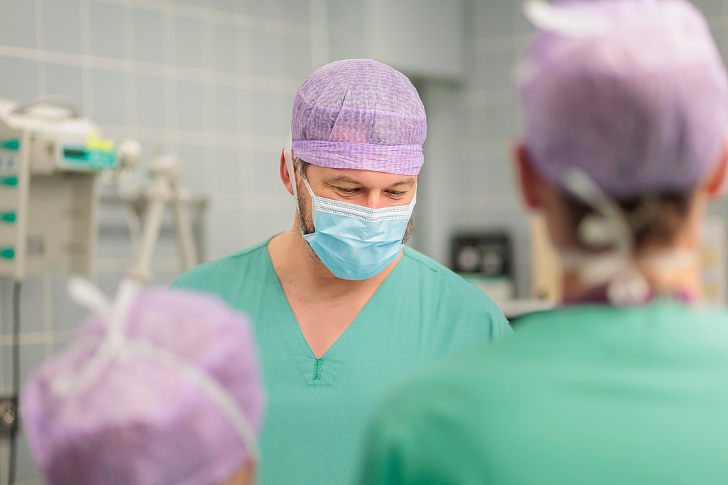 Drei medizinische Fachkräfte in grüner OP-Kleidung und lila Hauben in einem Krankenhausraum