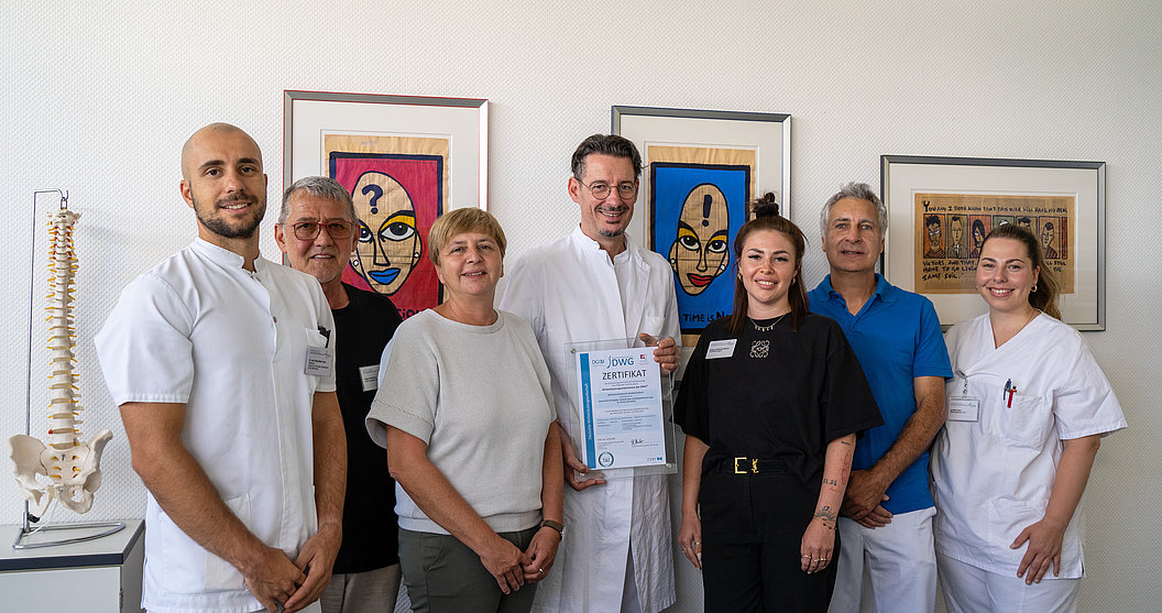 Gruppe von sieben Personen in medizinischer und Freizeitkleidung steht vor Wand mit farbigen Kunstwerken und einem Modell der menschlichen Wirbelsäule