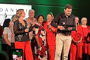 Eine Gruppe von Personen auf einer Bühne, einige tragen rote Kleidung. Eine Frau in schwarzer Bluse spricht ins Mikrofon, ein Mann in schwarzem Hemd hält ein Tablet. Im Hintergrund ist ein grünes Licht und ein Fan sichtbar.