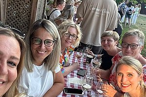 Gruppe von Menschen sitzt an einem langen Tisch mit rot-weiß kariertem Tischtuch, Wein- und Wassergläsern, Speisekarten und einer kleinen gelben Blume auf dem Tisch, draußen unter einem überdachten Bereich