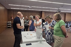 Sechs Personen stehen in einem Raum mit Holzwänden und diskutieren um zwei weiße Tische, auf denen Flaschen und ein Smartphone liegen.