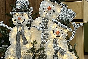 Drei beleuchtete Schneemannfiguren mit grauen Hüten und Schals, umgeben von Tannenzweigen und weißem Stoff.