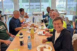 Mehrere Personen sitzen an einem langen Tisch in einem hellen Raum mit großen Fenstern und essen gemeinsam Mahlzeiten von Tellern.