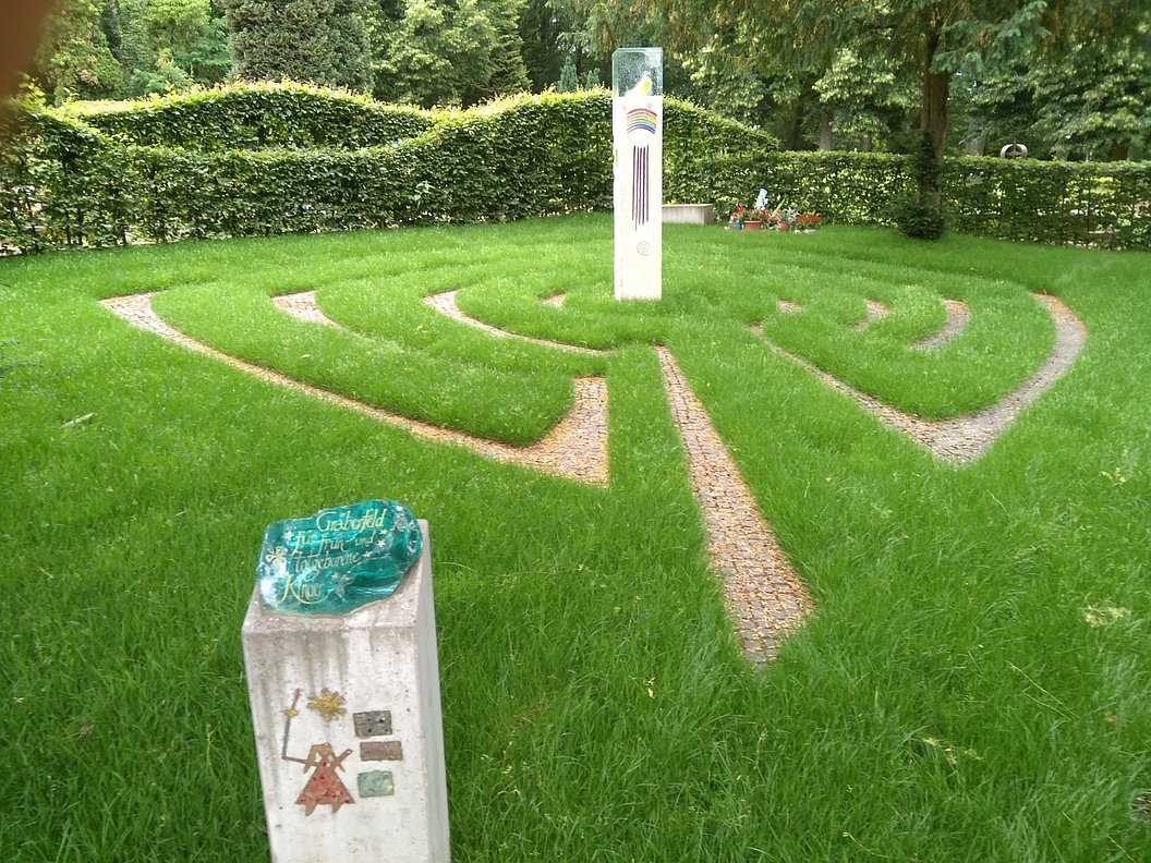 Grüner Rasen mit einem labyrinthartigen Muster aus gepflasterten Wegen und zwei weißen Steinsäulen, eine mit einer grünen Glasplatte und die andere mit einem vertikalen roten Muster
