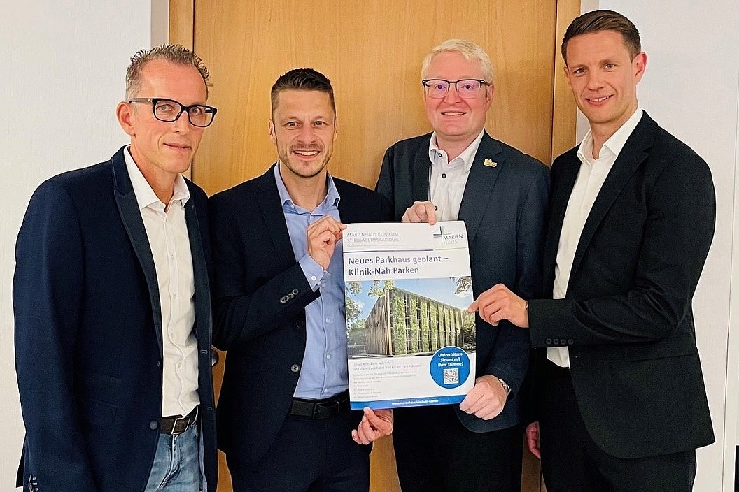 Vier Personen stehen zusammen und halten ein Plakat mit der Aufschrift 'Neues Parkhaus geplant - Klinik Nah Paken'. Im Hintergrund eine Holzverkleidung.