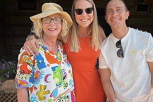 Drei Personen stehen zusammen, eine trägt ein buntes Hemd und Strohhut, eine andere ein rotes Kleid, die dritte ein weißes T-Shirt mit Sonnenbrille am Kragen
