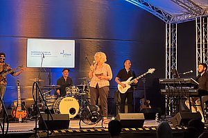 Eine Band tritt auf einer Bühne auf. Ein Sänger steht im Vordergrund, umgeben von Musikern an verschiedenen Instrumenten: Schlagzeug, E-Gitarre, E-Bass und Keyboard. Im Hintergrund ist ein Bildschirm mit Text.